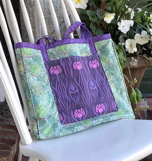 A hand made quilted book bag on a white chair with white flowers behind it.